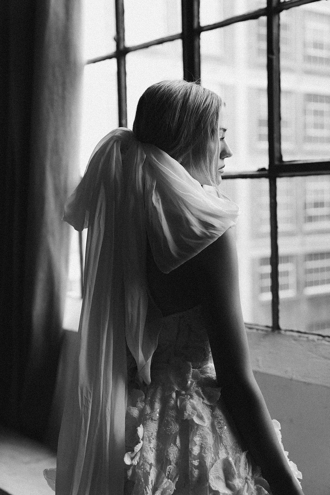 Black-and-white portrait of a bride in the Isla mini, wearing the detachable overskirt and sculpted veil, standing by industrial windows with soft natural light.